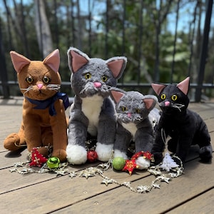 May include: Four plush cat toys in brown, grey, and black. Each cat has pink inner ears and a pink nose. The cats are decorated with Christmas ornaments and a star garland. The cats are sitting on a wooden surface.