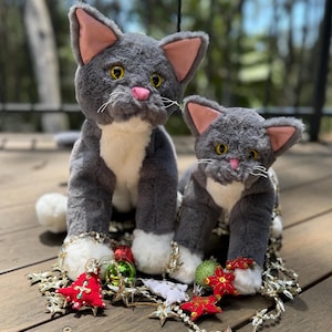 Peut inclure: Deux jouets en peluche en forme de chats gris avec des pattes blanches, des nez roses et des oreilles doublées de rose sont posés sur une surface en bois. Ils sont décorés d'ornements de Noël et d'une guirlande d'étoiles argentées. Les chats ont des yeux jaunes et des moustaches blanches.