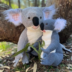 May include: Two grey plush koala bears, one larger than the other, are sitting together on a bed of brown leaves. The larger koala has pink and white checked ears, while the smaller koala has white fur around its ears. Both koalas have black noses and eyes.