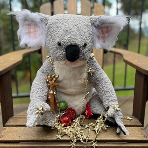 May include: A grey koala plush toy wearing a gold star garland and holding a small reindeer figurine. The koala is sitting on a wooden chair with red and green Christmas ornaments scattered around it.
