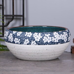 May include: A white ceramic planter with a blue and white floral design. The planter has a wide, round shape and a textured surface.