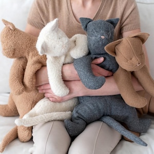 May include: Four plush cat toys in different colours and textures. One is light brown and fuzzy, one is cream-coloured and fuzzy, one is dark grey with a smooth fabric finish, and one is medium brown with a smooth fabric finish. Each cat has stitched facial features.