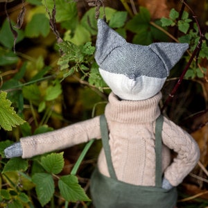 Puede incluir: Un zorro de peluche gris y blanco que lleva un suéter beige y un mono verde. El zorro está sentado en un lecho de hojas verdes.