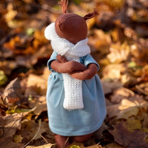 Könnte beinhalten: Eine braune Stoffpuppe mit einem weißen Strick Schal und einem blauen Kleid, das auf einem Bett aus Herbstlaub steht.