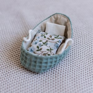 May include: A miniature teal cradle with a textured exterior, lined with cream-colored fabric. Inside, a small pillow and a rectangular pillow with a floral pattern of blue flowers and green leaves rest. A charming, handcrafted item.