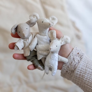 Może przedstawiać: Trzy małe, ręcznie robione lalki z tkaniny w kształcie myszy w dłoni. Myszki ubrane są w miniaturowe ubranka, w tym białą koszulę, sukienkę i spodnie. Lalki są głównie beżowe i białe, z małymi czarnymi oczami.