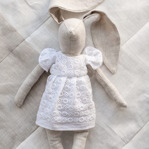 May include: A handmade linen bunny doll wearing a white eyelet dress. The doll has long ears and is lying on a white linen background.