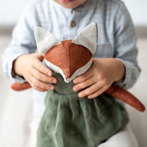 May include: A handmade fox doll with a brown head, white ears, and a green dress. The doll is held by a person wearing a light blue shirt. The fox doll is a soft toy.