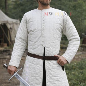 May include: A white, quilted medieval gambeson with long sleeves and a button-up front. The garment is worn with a brown belt. A silver sword is held in the hand. The image has the text "MW PLANET".