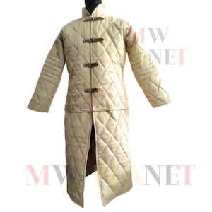 Op de afbeelding: Beige gewatteerde middeleeuwse gambeson met een opstaande kraag en lange mouwen. De gambeson heeft een ruitvormige stiksel en leren riemen om te sluiten. Dit historische kledingstuk is ontworpen voor bescherming en is geschikt voor re-enactments of kostuumevenementen.