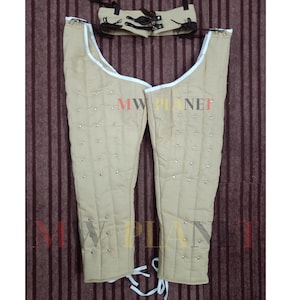 Può includere: Un paio di gambe in cuoio imbottite di colore beige con bordi bianchi e borchie metalliche. L'armatura è fissata con lacci bianchi. È inclusa una cintura in pelle marrone con lacci bianchi.
