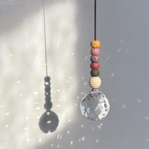 Könnte beinhalten: Ein Kristall-Sonnenfänger mit einer klaren Kristallkugel und einer Schnur mit bunten Perlen. Die Perlen sind gelb, rosa, grün und braun. Der Sonnenfänger hängt an einer schwarzen Schnur.