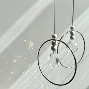 Könnte beinhalten: Zwei Kristall-Sonnenfänger hängen von der Decke. Jeder Sonnenfänger hat einen klaren Kristall-Tropfen, der an einem silbernen Ring mit weißen Perlen hängt.