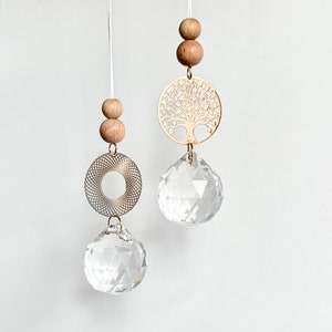 Könnte beinhalten: Zwei hängende Kristall-Sonnenfänger. Einer hat ein goldfarbenes Lebensbaum-Design, der andere ein kreisförmiges Filigran. Beide haben klare Kristallkugeln und Holzperlen, die an weißen Schnüren hängen.
