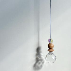 Könnte beinhalten: Ein Kristallprisma, das an einer weißen Schnur mit Holzperlen hängt. Das Prisma ist klar und hat ein facettierendes Design. Die Perlen sind braun und haben eine natürliche Holzoptik.