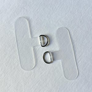 Könnte beinhalten: Zwei durchsichtige, T-förmige Acryl-Laschen, die jeweils mit einem kleinen, silberfarbenen D-Ring aus Metall am mittleren Steg versehen sind. Die Laschen haben abgerundete Kanten und eine glatte, transparente Oberfläche zur Befestigung.