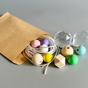 Könnte beinhalten: Ein brauner Papierumschlag mit einer weißen Schnur und verschiedenen Perlen darin. Die Perlen sind in Pastellfarben, darunter rosa, lila, gelb und grün. Es gibt auch zwei klare Kristallkugeln.