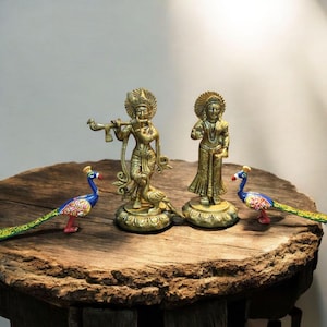 May include: A collection of brass religious statues and decorative peacocks on a wooden table. The statues depict figures with ornate headdresses and clothing. The peacocks are painted with vibrant colors, including blue, green, and red.