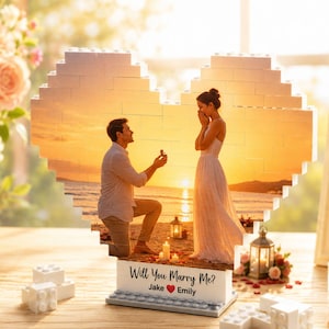 Könnte beinhalten: Ein herzförmiges Baustein-Display mit einem aufgedruckten Bild eines Heiratsantrags am Strand bei Sonnenuntergang. Der Sockel trägt den Text "Will You Marry Me?" sowie die Namen "Jake" und "Emily" mit einem roten Herzsymbol.