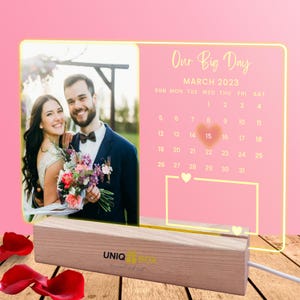 May include: A personalized wedding photo frame with a photo of a smiling couple. The frame includes a calendar for March 2023 with the text "Our Big Day". The base is made of wood and has the brand name "UNIQ".