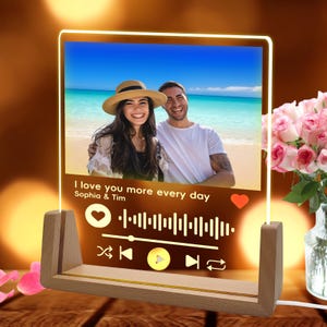 Może przedstawiać: Spersonalizowana lampka nocna LED ze zdjęciem pary na plaży. Światło ma drewnianą podstawę i wyświetla tekst "I love you more every day" z interfejsem odtwarzacza muzyki. Światło emituje ciepłą, zachęcającą poświatę.