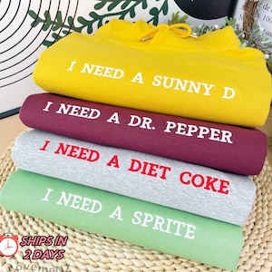 May include: A stack of four hoodies with different colors and slogans. The top hoodie is yellow with the text "I NEED A SUNNY D". The second hoodie is maroon with the text "I NEED A DR. PEPPER". The third hoodie is gray with the text "I NEED A DIET COKE". The bottom hoodie is green with the text "I NEED A SPRITE".