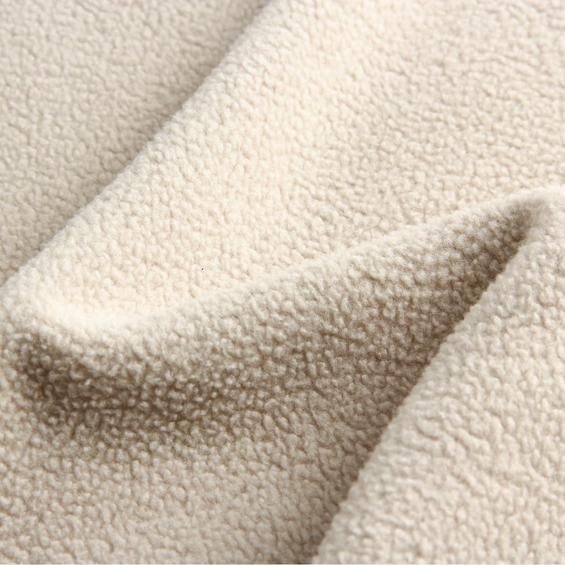 Puede incluir: Primer plano de una tela suave de color crema con una textura peluda. La tela est&aacute; hecha de un material afelpado que es perfecto para una manta o una manta acogedora.