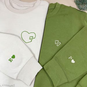 以下が含まれることがあります： 刺繍デザインの白いスウェットシャツと緑のスウェットシャツ2枚。白いスウェットシャツには、パズルのピースが付いた緑色のハートと、ハートの文字「E」があります。緑色のスウェットシャツには、パズルのピースとハートの文字「P」があります。