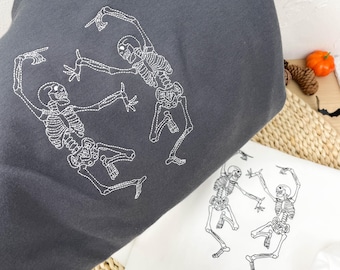 May include: Dark gray sweatshirt and white sweatshirt featuring a white embroidered design of two dancing skeletons. The sweatshirts are folded, showcasing the Halloween-themed graphic. The brand name "LovemeNZ" is visible in the top left corner.