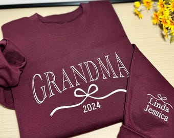Könnte beinhalten: Burgunderfarbener Langarm-Sweatshirt mit dem Text "Grandma" in Weiß mit einem weißen Band und einer Schleife darunter. Das Jahr 2024 ist unter der Schleife gedruckt. Der rechte Ärmel trägt den Text "Linda Jessica" in Weiß mit einer weißen Schleife.
