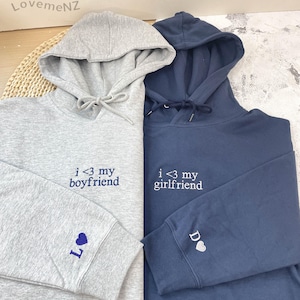 Könnte beinhalten: Zwei Kapuzenpullover, einer grau und einer marineblau. Der graue Pullover hat "i <3 my boyfriend" auf der Vorderseite und ein Herz und den Buchstaben "L" auf dem Ärmel gestickt. Der marineblaue Pullover hat "i <3 my girlfriend" auf der Vorderseite und ein Herz und den Buchstaben "D" auf dem Ärmel gestickt.
