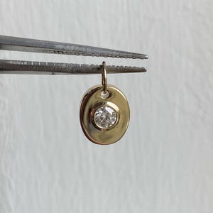 Op de afbeelding: Een gouden hanger met een heldere, ronde edelsteen in het midden. De ovale hanger wordt vastgehouden door een pincet. Het heeft een kleine lus aan de bovenkant voor een ketting.