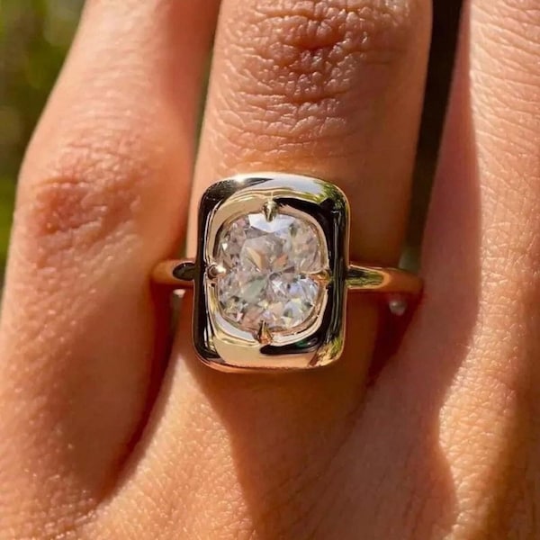 May include: A gold ring featuring a large, oval-shaped diamond set in a bezel setting. The diamond is surrounded by a gold frame.
