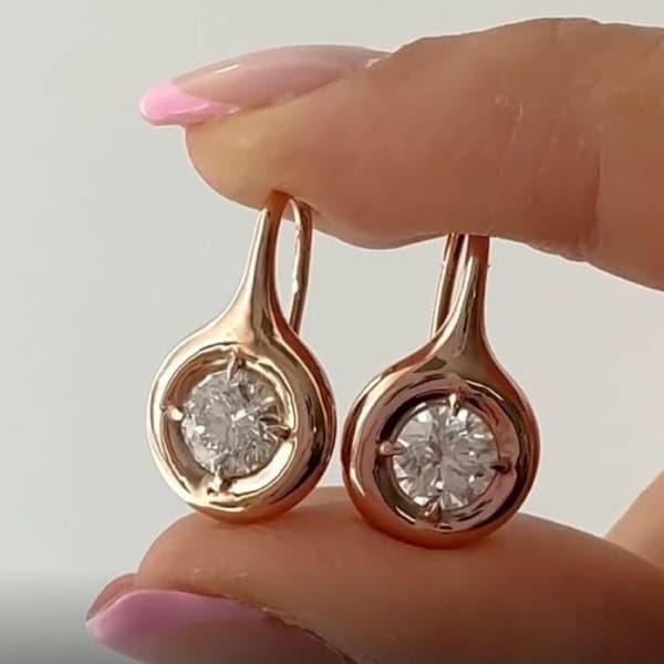 May include: A pair of rose gold earrings with a teardrop design. Each earring features a round, clear gemstone set in a bezel. The earrings are held up against a light background.