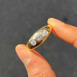 Puede incluir: Un anillo de oro con tres piedras preciosas redondas y transparentes dispuestas en fila. El anillo tiene una superficie lisa y pulida y se sostiene entre dos dedos sobre un fondo oscuro. Las piedras preciosas brillan a la luz.