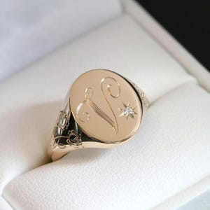 Könnte beinhalten: Goldener Siegelring mit ovaler Oberfläche. Die Oberfläche zeigt einen gravierten Buchstaben "V" und ein Sternmuster mit einem kleinen Diamanten. Der Ring hat florale Details am Band.