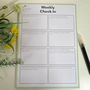 May include: A weekly check-in sheet with a pale green border and the text "Weekly Check In." The sheet includes prompts for self-reflection, such as "What has gone well this week?" and "What is my goal or homework for this week?" A black pen rests on the right.