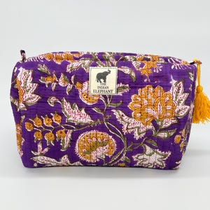 May include: A quilted purple pouch featuring a floral pattern in yellow, white, and green. A rectangular label on the front displays an elephant icon and the text THE INDIAN ELEPHANT. A yellow tassel is attached to the side zipper pull.
