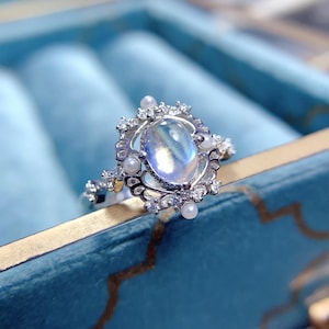 Puede incluir: Un anillo de plata con una gran piedra de luna ovalada engastada en un delicado diseño de filigrana. El anillo está adornado con pequeñas perlas blancas y piedras de circonita cúbica transparentes.