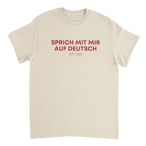 May include: A cream-colored t-shirt with the German phrase "SPRICH MIT MIR AUF DEUTSCH ich übe" printed in red. The text translates to "Speak to me in German, I am practicing."