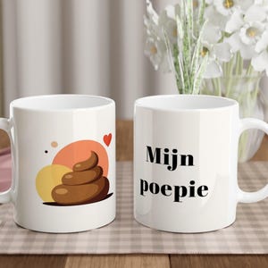 Pode incluir: Duas canecas de cerâmica brancas. Uma caneca apresenta um emoji de cocó castanho com círculos laranja e amarelos e um coração vermelho. A outra caneca tem as palavras "Mijn poepie" em preto. Ambas as canecas têm uma alça branca.