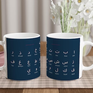 Puede incluir: Dos tazas de cerámica blanca con fondo azul oscuro. Las tazas están decoradas con letras árabes blancas. Las tazas están sobre una mesa con un paño rosa y utensilios de madera. Flores blancas en un jarrón al fondo.