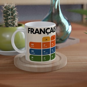 Op de afbeelding: Een witte keramische mok met het woord FRANÇAIS in dikke zwarte letters bovenaan. Onder de tekst toont een raster Franse werkwoordvervoegingspatronen in oranje, blauw en groen kleurgecodeerde secties met witte tekst voor voornaamwoorden en uitgangen.
