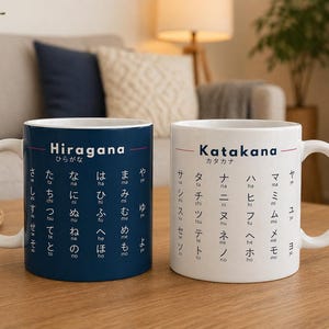 以下が含まれることがあります： 日本語のひらがなとカタカナの文字がプリントされた白いセラミックマグカップが2つ。1つのマグカップは濃い青色、もう1つは白色です。マグカップは木製のテーブルの上にあります。