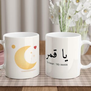 Puede incluir: Dos tazas de cerámica blanca con asas. Una taza presenta un diseño de media luna con un corazón y estrellas. La otra taza tiene texto árabe y la traducción al inglés "My moon". Ambas tazas están sobre una superficie a cuadros.