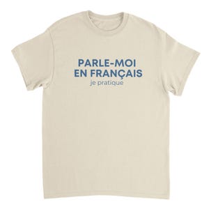 以下が含まれることがあります： クリーム色のTシャツに青い文字で「PARLE-MOI EN FRANÇAIS je pratique」と書かれています。Tシャツはクルーネックで半袖です。テキストは「フランス語で話しかけてください、練習します」と翻訳されます。