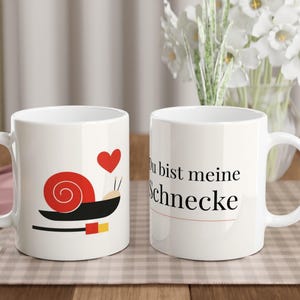 Könnte beinhalten: Zwei weiße Keramikbecher. Ein Becher zeigt ein rotes Schnecken-Design mit einem Herz, der andere sagt "Du bist meine Schnecke" in schwarzem Text. Beide Becher haben einen weißen Griff und stehen auf einer karierten Oberfläche.