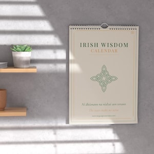 Könnte beinhalten: Ein cremefarbenes Wandkalender mit dem Titel "Irish Wisdom Calendar" mit grünem Text und einem keltischen Knotenmuster. Der Kalender hängt an einer grauen Wand, mit einer kleinen Pflanze und anderen Gegenständen auf Holzregalen.