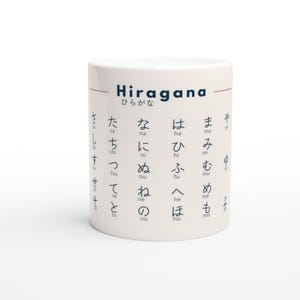 May include: White ceramic mug with the word "Hiragana" in navy blue, along with Japanese characters. The mug features a red horizontal line near the top and is printed with Japanese characters in a grid pattern.