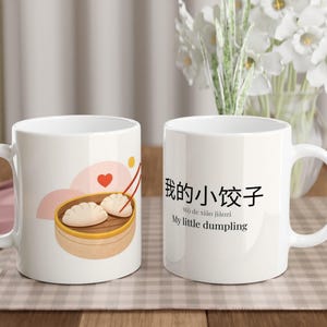 May include: Two white ceramic mugs. One mug features a dumpling illustration with a heart and chopsticks. The other mug has Chinese text and the English phrase "My little dumpling". Both mugs have a curved handle.